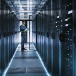 IT Technician Works on Laptop in Big Data Center full of Rack Servers. He Runs Diagnostics and Maintenance, Sets System Up.