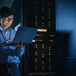 In Dark Data Center: Male IT Specialist Stands Beside the Row of Operational Server Racks, Uses Laptop for Maintenance. Concept for Cloud Computing, Artificial Intelligence, Supercomputer, Cybersecurity. Neon Lights